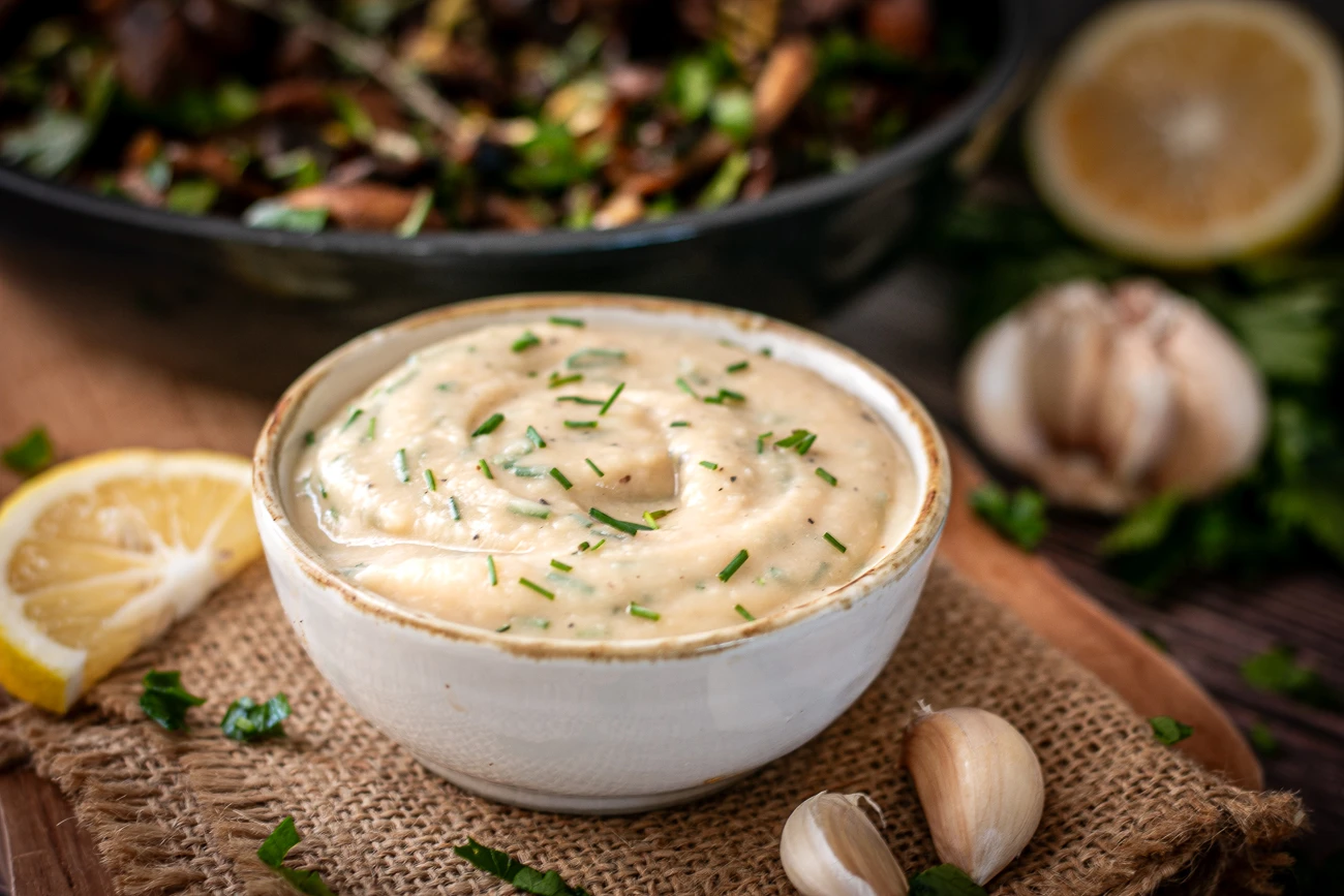 Schale mit veganem Knoblauch-Dip vor einer Pfanne mit gebratenen Champignons, Austernpilzen und Petersilie