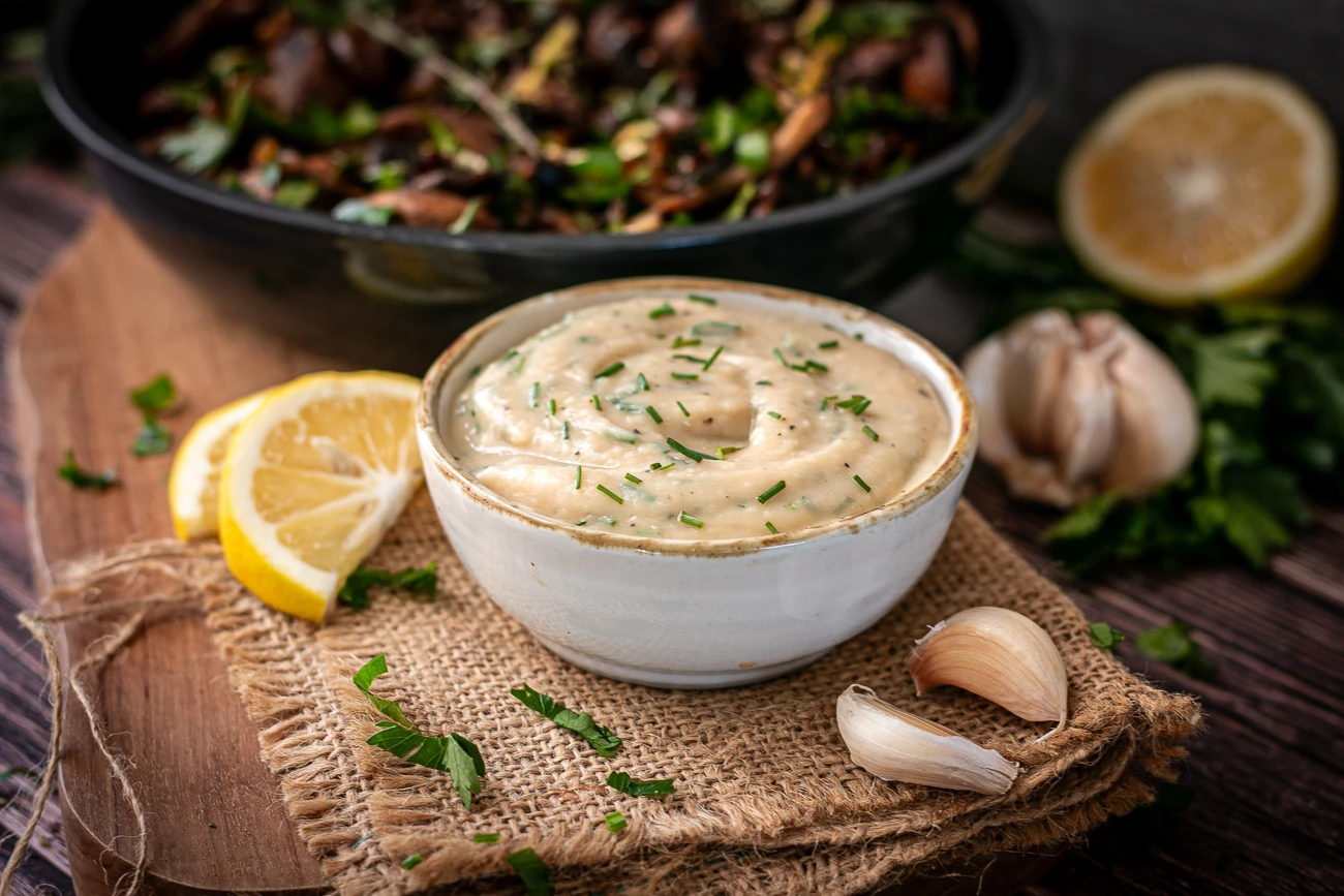 Cremiger Knoblauch-Dip aus weiĂźen Bohnen auf einem Holzbrett mit Jutetuch, Zitrone, Knoblauch und frischer Petersilie