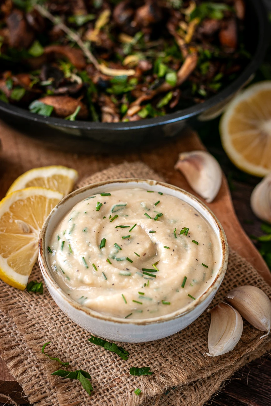 Selbstgemachter Knoblauch-Dip ohne Mayo mit Schnittlauch und Petersilie, daneben Knoblauchzehen und Zitrone