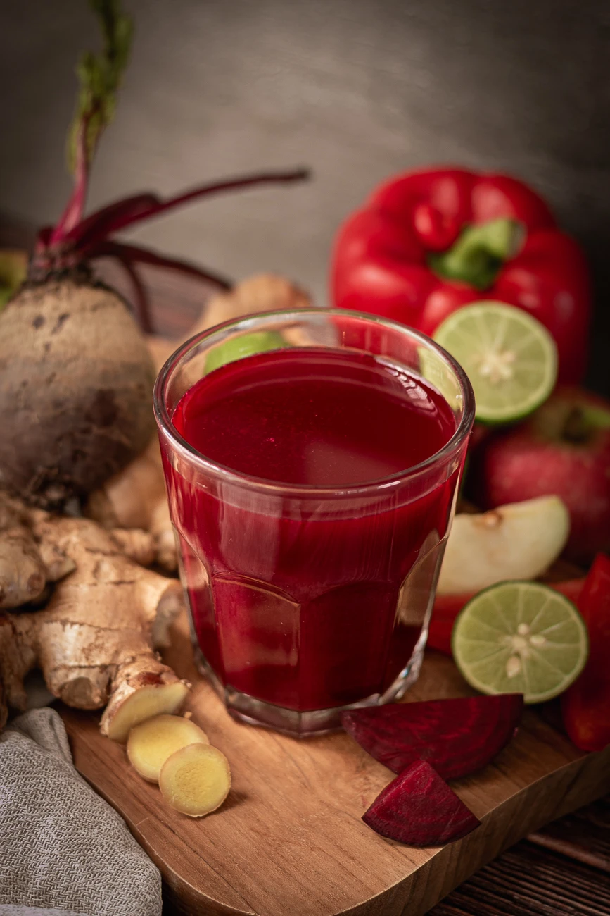Ein Glas Rote Bete Saft, daneben Zutaten wie Rote Bete, rote Paprika, Limette, Äpfel und Ingwer