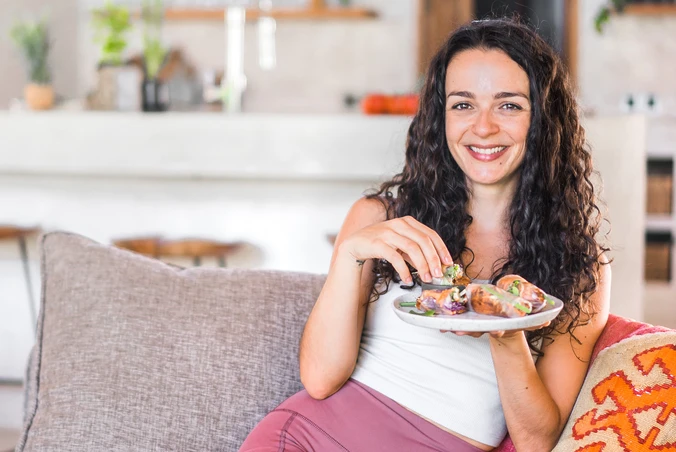 Sofia Konstantinidou, vegane Ernährungsberaterin, sitzt lächelnd auf einem Sofa und isst Sommerrollen